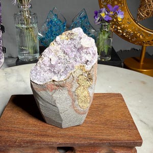 May include: A raw amethyst geode specimen featuring a cluster of light purple crystals set within a rough, grey and tan stone matrix. The geode has a jagged, uneven top edge and a solid, textured exterior with visible mineral deposits.