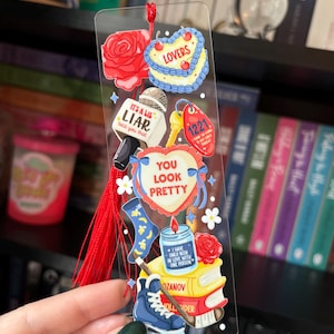 May include: Clear bookmark with various colorful illustrations. Designs include a red rose, a heart-shaped cake, a microphone with the text "IT'S A LIE LIAR told you that", a key tag with "1221", and a heart with "YOU LOOK PRETTY".