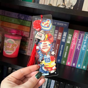 May include: A clear acrylic bookmark with various colorful charms and a red tassel. Charms include a rose, a heart-shaped cake, a microphone with "It's a LIAR", a key tag with "1221", and a blue ice skate. The text "YOU LOOK PRETTY" is also visible.