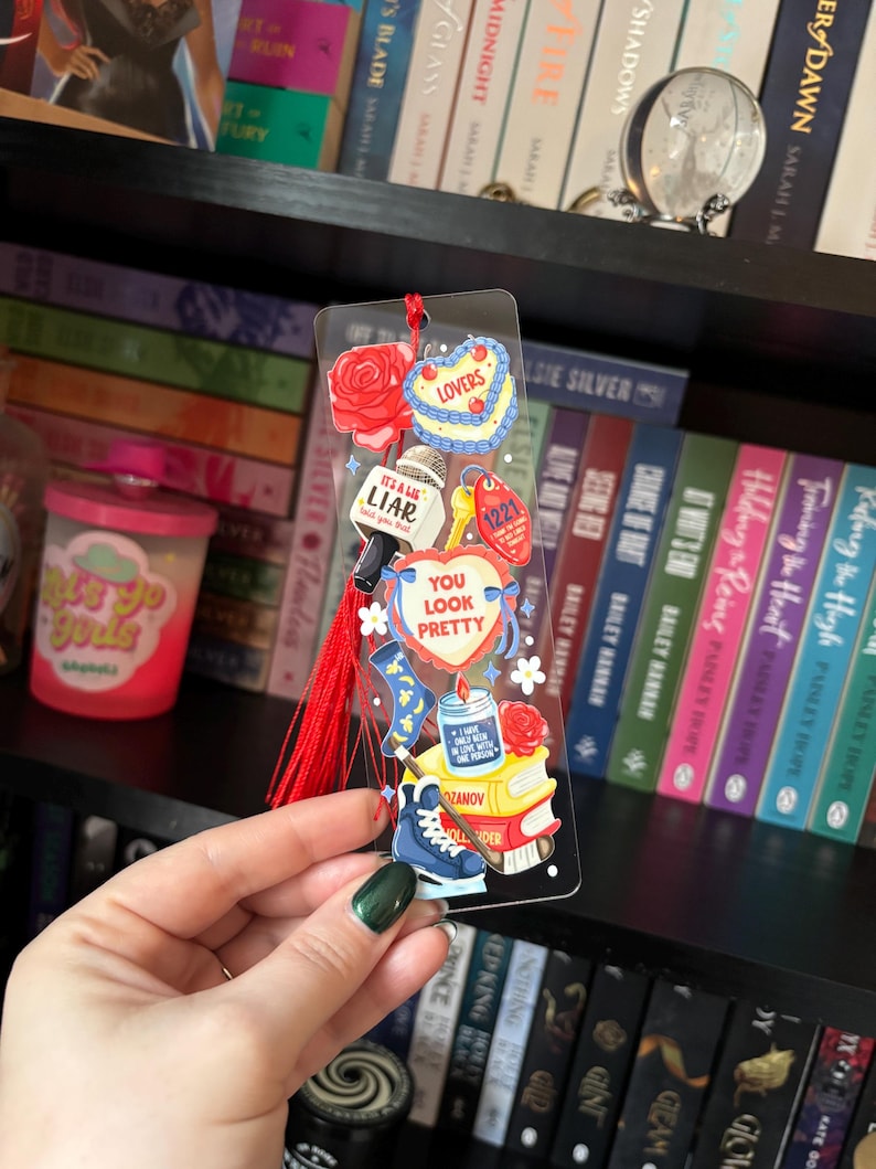 May include: A clear acrylic bookmark with various colorful charms and a red tassel. Charms include a rose, a heart-shaped cake, a microphone with "It's a LIAR", a key tag with "1221", and a blue ice skate. The text "YOU LOOK PRETTY" is also visible.