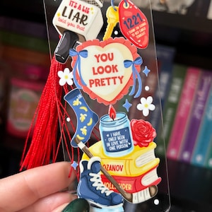 May include: A clear bookmark with various illustrations and text. The bookmark features a microphone, a heart with the words "YOU LOOK PRETTY", a key tag with "1221", a candle, a rose, a sock, and a hockey skate. A red tassel hangs from the top.