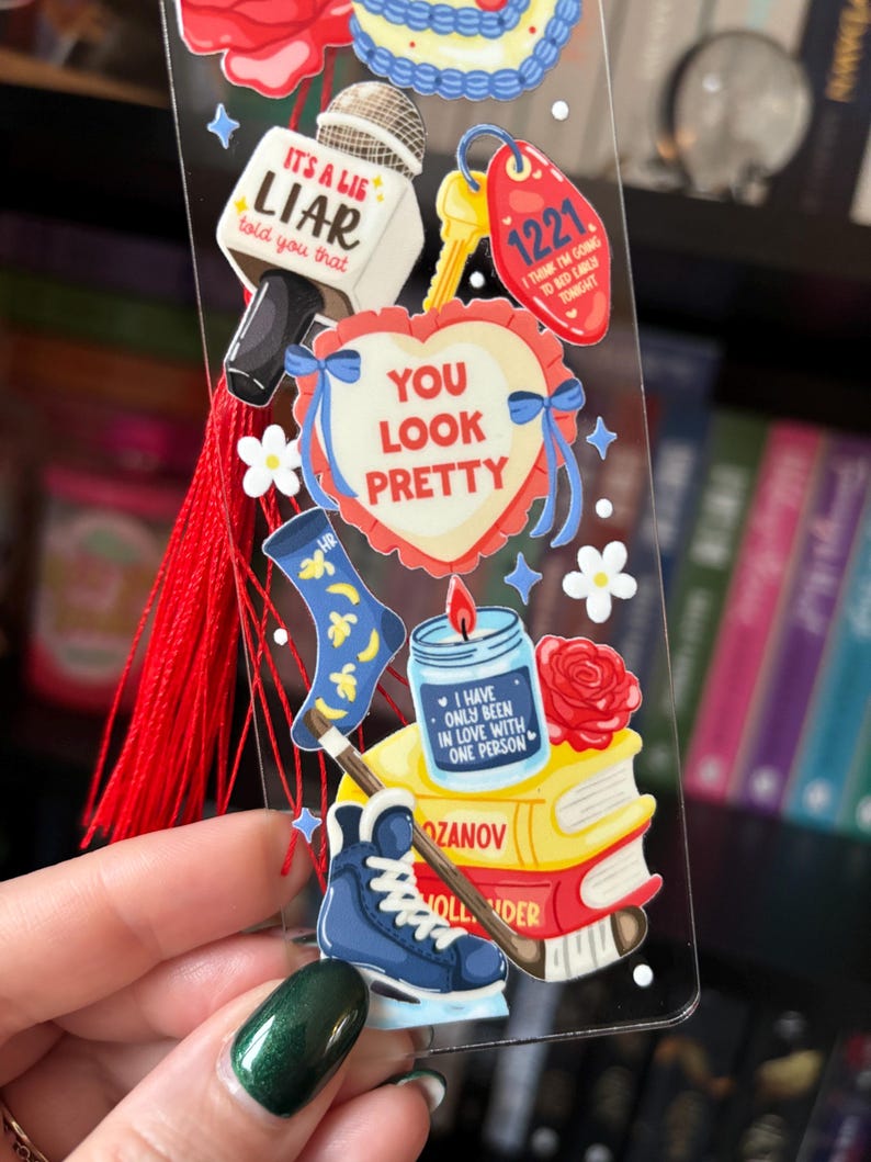 May include: A clear bookmark with various illustrations and text. The bookmark features a microphone, a heart with the words "YOU LOOK PRETTY", a key tag with "1221", a candle, a rose, a sock, and a hockey skate. A red tassel hangs from the top.