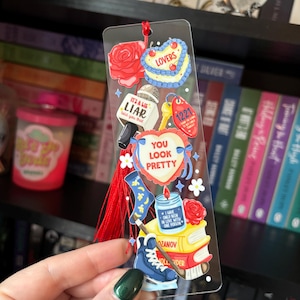 May include: A clear acrylic bookmark with various colorful illustrations, including a rose, a cake that says "LOVERS", a microphone with the text "IT'S A LIE LIAR told you that", and a heart that says "YOU LOOK PRETTY". A red tassel hangs from the top.