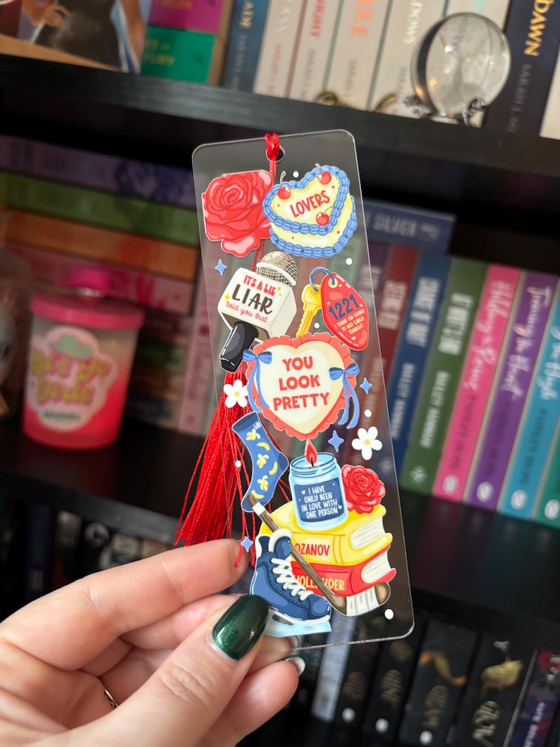 May include: A clear acrylic bookmark with various colorful illustrations, including a rose, a cake that says "LOVERS", a microphone with the text "IT'S A LIE LIAR told you that", and a heart that says "YOU LOOK PRETTY". A red tassel hangs from the top.
