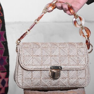 May include: A beige woven handbag with a brown and clear plastic chain strap. The bag has a gold clasp and a geometric pattern.
