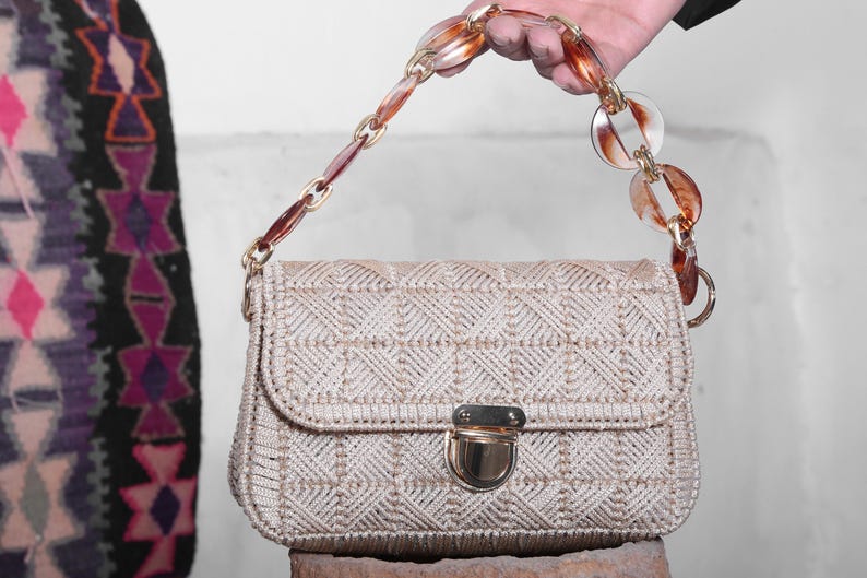 May include: A beige woven handbag with a brown and clear plastic chain strap. The bag has a gold clasp and a geometric pattern.