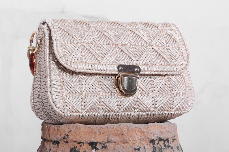 May include: A white woven crossbody bag with a gold clasp closure. The bag has a geometric pattern and is made of a thick, textured material.
