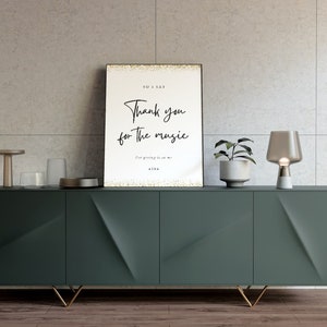 Puede incluir: Una impresión enmarcada blanca con detalles de purpurina dorada y el texto "Yo I Say Thank you for the music I'm giving it to me ABBA". La impresión está sobre un gabinete verde oscuro con patas doradas. Hay una pequeña planta en maceta y una lámpara sobre el gabinete.