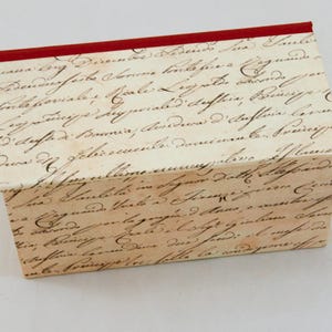 Peut inclure: Une boîte rectangulaire avec un couvercle rouge et des côtés recouverts d'une écriture manuscrite délavée. La boîte a une esthétique vintage, le texte apparaissant dans une teinte sépia, suggérant un thème antique ou historique.