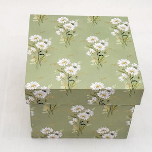 Può includere: Una scatola regalo quadrata con coperchio, con un motivo ripetuto di margherite bianche con centri gialli e steli verdi su sfondo verde chiaro. La scatola è di cartone.