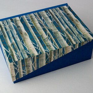 Op de afbeelding: Een blauwe decoratieve doos met een deksel met een berkenboomontwerp. Het deksel heeft een patroon van witte berkenstammen tegen een teal achtergrond met kleine oranje accenten. De zijkanten van de doos zijn effen blauw.