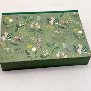 Könnte beinhalten: Eine rechteckige grüne Schachtel mit einem floralen Muster auf dem Deckel. Das Muster zeigt verschiedene Blumen und Blätter in Rosa, Weiß, Gelb und Blau auf grünem Hintergrund. Die Schachtel hat einen einfarbigen grünen Boden und einen grünen Rand.