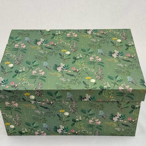 Op de afbeelding: Een rechthoekige opbergdoos met een groen bloemenpatroon. De doos heeft een herhalend ontwerp van verschillende bloemen en bladeren in tinten wit, roze, geel en blauw tegen een groene achtergrond. De doos is gesloten en staat op een wit oppervlak.