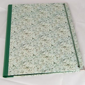 Könnte beinhalten: Ein Ordner mit floralem Muster mit grünem Rücken und elastischem Verschluss. Der Einband hat einen hellblauen Hintergrund mit einem sich wiederholenden Muster aus weißen Blumen und grünen Blättern. Der Ordner hat einen Eckenschutz aus Metall.