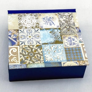 Könnte beinhalten: Eine quadratische Box mit marineblauem Boden und einem Deckel, der mit einem Patchwork aus gemusterten Fliesen verziert ist. Die Fliesen weisen florale und geometrische Designs in Blau-, Beige- und Goldtönen auf. Die Box eignet sich zum Aufbewahren oder Verschenken.