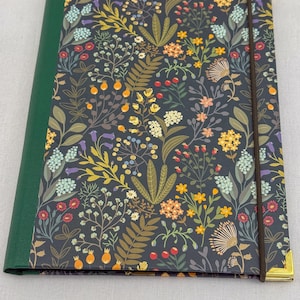 May include: A floral journal with a dark background and a green spine. The cover features a pattern of colorful flowers and foliage, including yellow, orange, and blue blooms. A brown elastic band and gold corner protectors add detail.