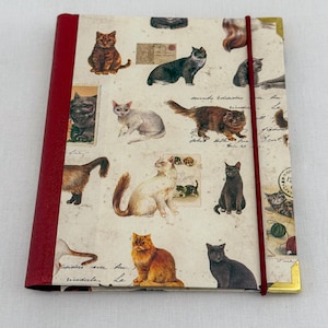 May include: A journal with a cream cover adorned with illustrations of cats in various poses. It has a red spine and a red elastic closure. The corners are finished with gold detailing.