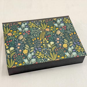 Peut inclure: Boîte rectangulaire avec un couvercle à motifs floraux vert foncé. Le couvercle présente un motif détaillé de diverses fleurs et feuillages dans des tons de jaune, orange, rose et bleu. La boîte a une base noire unie.
