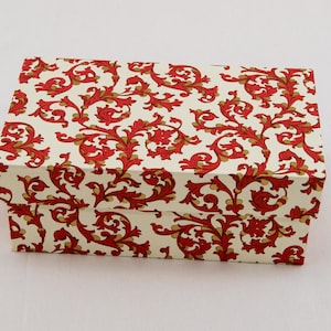 Op de afbeelding: Een rechthoekige doos met deksel, versierd met een crèmekleurige achtergrond en een herhalend patroon van rode en gouden gestileerde bloemen- en krulontwerpen. Geschikt voor cadeau geven of opslag.