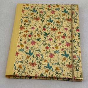 Puede incluir: Un cuaderno con diseño floral y cubierta amarilla. El patrón incluye flores rojas, rosas y amarillas con hojas verdes sobre un fondo crema. Tiene una banda elástica beige y protectores de esquina dorados. El cuaderno mide aproximadamente 20 cm.