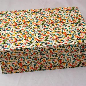 Può includere: Una scatola rettangolare con coperchio, ricoperta da un motivo floreale colorato. Il design presenta uno sfondo crema con un motivo ripetuto di fiori e foglie stilizzate rosse, gialle, blu e verdi. La scatola è per la conservazione o l'imballaggio regalo.