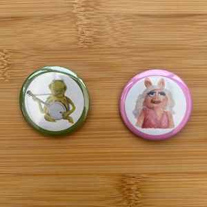 Op de afbeelding: Twee ronde buttons met Muppet-personages. Eén button heeft Kermit de Kikker die banjo speelt, omringd door een groene rand. De andere button toont Miss Piggy met een roze rand. Beide buttons liggen op een houten oppervlak.
