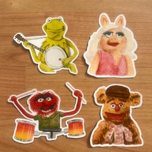 Peut inclure: Quatre autocollants colorés représentant des personnages des Muppets. Kermit la Grenouille joue du banjo, Miss Piggy porte une robe rose, Animal joue de la batterie et Fozzie Bear porte un chapeau et une chemise à motifs. Les autocollants sont sur une surface en bois.