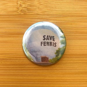 May include: Round button with a graphic of a cloudy sky, a building, and the text "SAVE FERRIS" in black. The button has a glossy finish and is set against a wooden background.