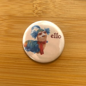 Könnte beinhalten: Ein runder Button mit einer farbenfrohen Illustration eines wurmartigen Wesens mit blauen, orangefarbenen und roten Akzenten. Das Wort "ello" ist in dunkler roter Schrift auf der rechten Seite des Buttons aufgedruckt.