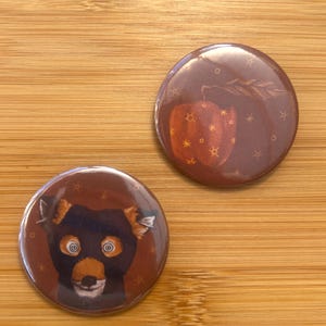 Könnte beinhalten: Zwei runde Buttons mit braunem Hintergrund. Ein Button zeigt ein bärenähnliches Wesen mit orangefarbenem und schwarzem Fell und großen Augen. Der andere Button hat einen braunen Apfel mit sternförmigen Akzenten.