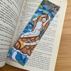 Könnte beinhalten: Ein farbenfrohes Lesezeichen mit einer Buntglas-Illustration einer Frau mit langen blonden Haaren. Das rechteckige Lesezeichen befindet sich in einem geöffneten Buch.