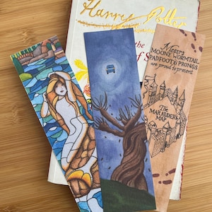 May include: A stack of three illustrated bookmarks and a hardcover book. The bookmarks feature a mermaid, a car in the night sky, and a map with text. The book has gold lettering that reads "Harry Potter".