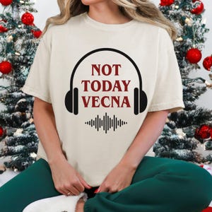May include: Cream-colored t-shirt with the text "NOT TODAY VECNA" in red and black, framed by a headphone design. Below the text is a black sound wave graphic. The shirt is short-sleeved.