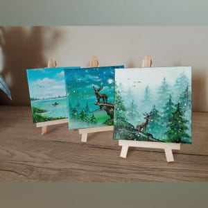 Può includere: Tre piccoli dipinti di paesaggi su cavalletti di legno. I dipinti raffigurano scene naturali serene, tra cui un lago, una notte stellata con un cervo e una foresta innevata con cervi. La tavolozza dei colori comprende blu, verdi e bianchi.