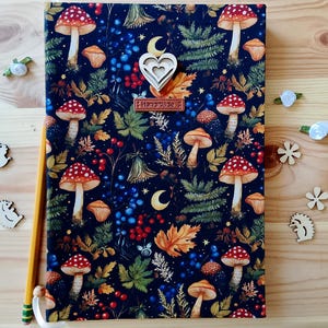 Può includere: Un taccuino rettangolare con copertina in tessuto blu scuro caratterizzato da un motivo di funghi rossi e bianchi, felci verdi, foglie autunnali e bacche. Sono presenti un ciondolo in legno a forma di cuore e un'etichetta in pelle Handmade.