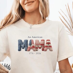 Peut inclure: Un t-shirt à manches courtes de couleur crème avec le texte An American MAMA 1776 - 2026. Le mot MAMA est imprimé dans une grande police gonflée en trois dimensions avec un motif patchwork rouge, blanc et bleu rappelant le drapeau américain.