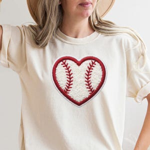 Puede incluir: Camiseta color crema con un diseño de corazón de béisbol. El corazón está delineado en rojo con un centro blanco texturizado y detalles de costura rojos, creando un look deportivo. La camiseta tiene mangas cortas y un corte relajado.