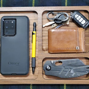 May include: A wooden tray with four compartments. The compartments contain a black phone case with the OtterBox logo, a yellow pen, a brown leather wallet with the text "www.bellroy.com" and a black pocket knife.