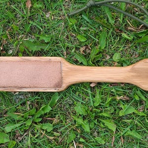 Puede incluir: Un strop de cuero sobre madera con una superficie de cuero marrón y un agujero en el mango. El strop está sobre un lecho de hierba verde.