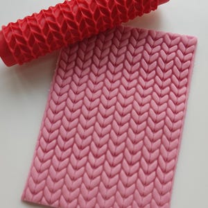 May include: A red textured rolling pin and a pink rectangular mat, both with a repeating knit pattern. The rolling pin is cylindrical with a handle on each end. The mat has a raised, textured surface.