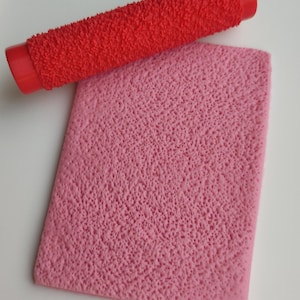 May include: A red textured rolling pin rests above a pink textured rectangular mat. The rolling pin has a cylindrical shape with a textured surface. The pink mat has a rectangular shape with a textured surface.