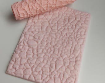 Puede incluir: Un rodillo texturizado rosa claro y una esterilla rectangular a juego. El rodillo es cilíndrico y tiene un patrón irregular en relieve. La esterilla tiene un patrón similar, formando un conjunto. Diseñado para manualidades o repostería.