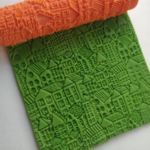 Puede incluir: Un rodillo naranja con un detallado patrón de paisaje urbano descansa sobre una superficie verde texturizada con un diseño de paisaje urbano a juego. El rodillo es cilíndrico y presenta un patrón repetitivo de edificios.