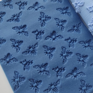 May include: A blue rolling pin and a blue textured sheet with a bee pattern. The rolling pin is cylindrical with a bee pattern. The sheet has a repeating pattern of bees pressed into the surface.