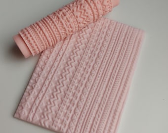 Puede incluir: Un rodillo texturizado de color rosa claro y una esterilla rectangular a juego. El rodillo es cilíndrico con un patrón repetitivo. La esterilla tiene varios patrones texturizados, incluyendo zigzags y líneas verticales. Los artículos son probablemente para hornear o manualidades.