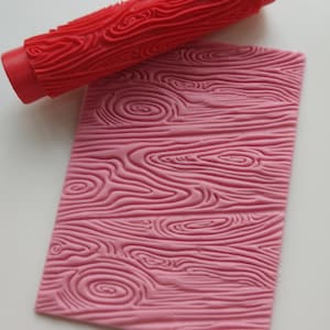 May include: A red and pink silicone rolling pin and mat with a wood grain pattern. The rolling pin is used to create a wood grain texture on fondant or other edible materials.