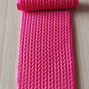 Puede incluir: Un rodillo de silicona rosa con una superficie texturizada que se asemeja a un patrón de punto. El rodillo está sobre una lámina plana de silicona rosa con el mismo patrón de punto.