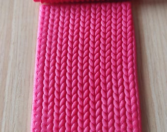 Puede incluir: Un rodillo de amasar rosa brillante y una esterilla rectangular a juego, ambos con un patrón de punto de cable en relieve. El rodillo es cilíndrico y la esterilla es rectangular. Los artículos son probablemente para hornear o manualidades.