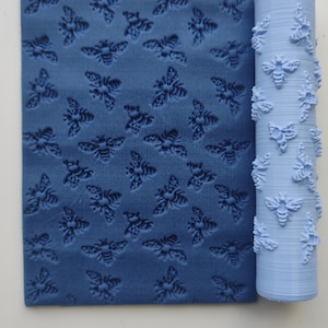 May include: A blue rolling pin and a matching textured sheet, both featuring a repeating bee pattern. The rolling pin is cylindrical, and the sheet is rectangular. The bee design is embossed on both items.