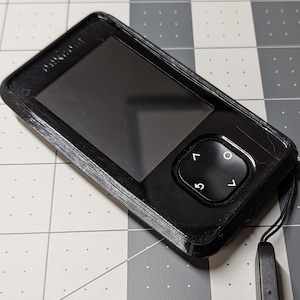 以下が含まれることがあります： 画面とボタンを備えた黒い電子デバイスで、黒い保護ケースに入っています。リストストラップが取り付けられています。デバイスは、グレーと白のチェック柄の表面にあります。